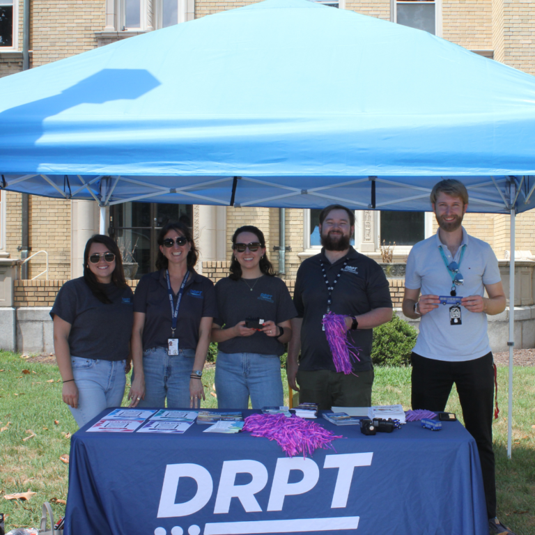 DRPT स्टाफ नीले रंग के टेंट के नीचे DRPT ब्रांडेड टेबल पर प्रमोशनल आइटम पकड़े हुए